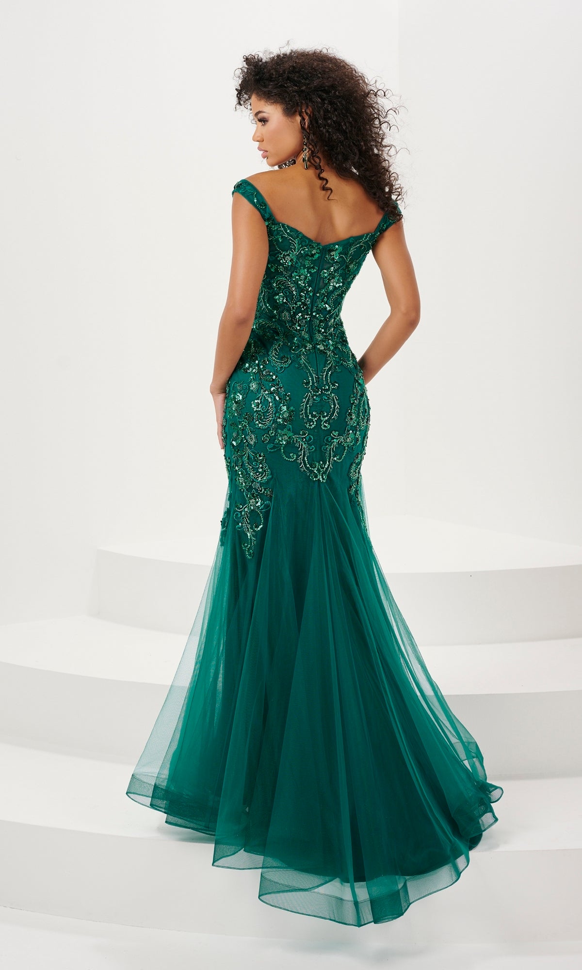 A woman stands on white steps, elegantly viewed from the back in the Long Prom Dress 14185 by Panoply—an off-the-shoulder, floor-length green mermaid gown with lace and sequin embroidered detailing.